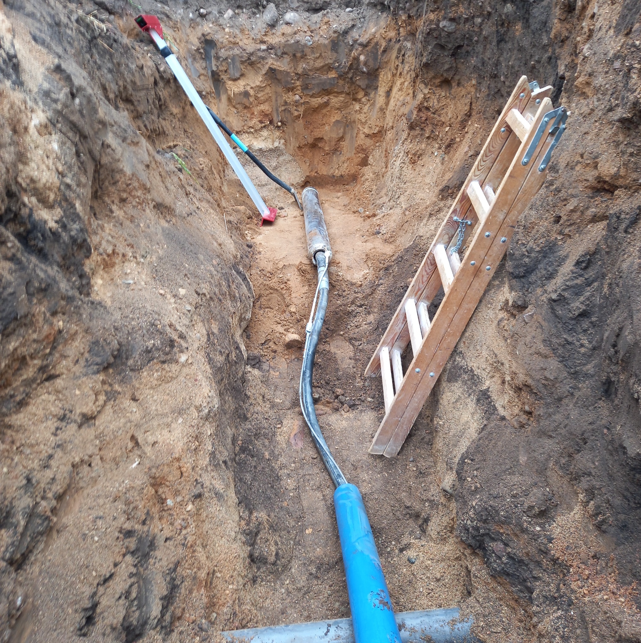 Wykop z rurą w ziemi, widoczna drabina i narzędzia. Prace ziemne związane z układaniem rur w wykopie. Rura z niebieskim zakończeniem.