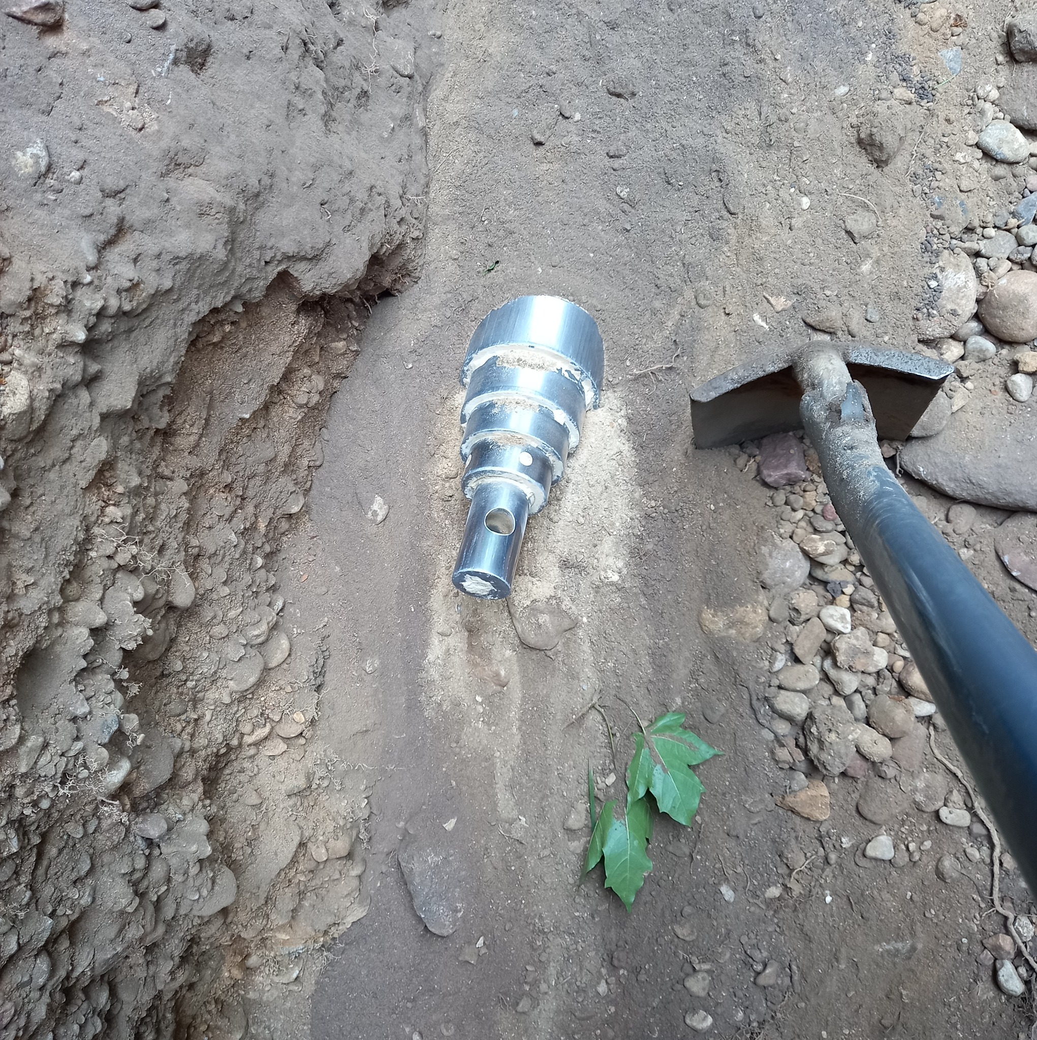 Metalowa rura w wykopie ziemnym, obok narzędzie. Widoczne świeże liście. Prace ziemne przyłącza wod-kan.