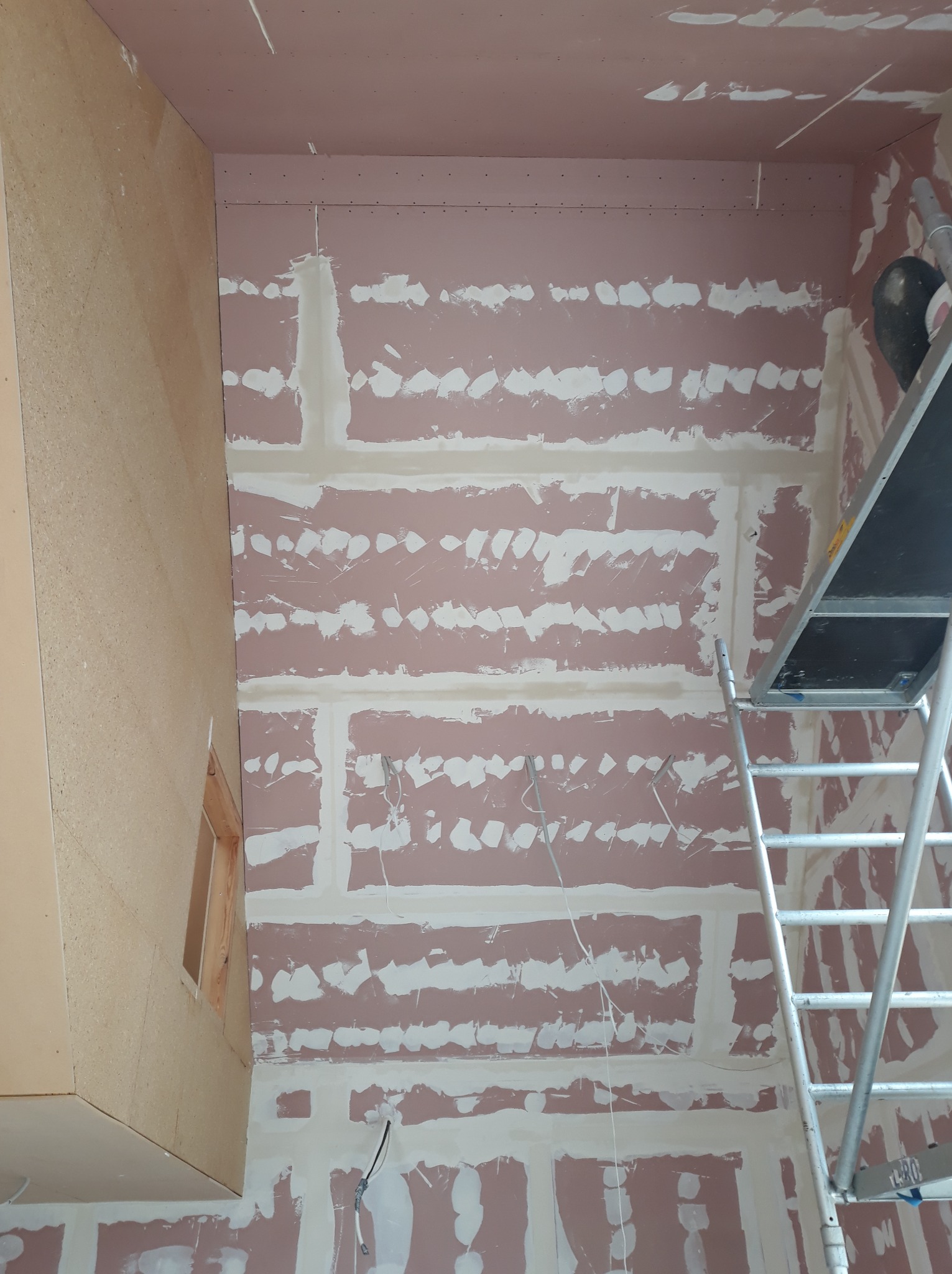 Ściana z płyt gipsowo-kartonowych w trakcie szpachlowania, widoczna drabina i wystające kable. Prace wykończeniowe wewnątrz budynku.