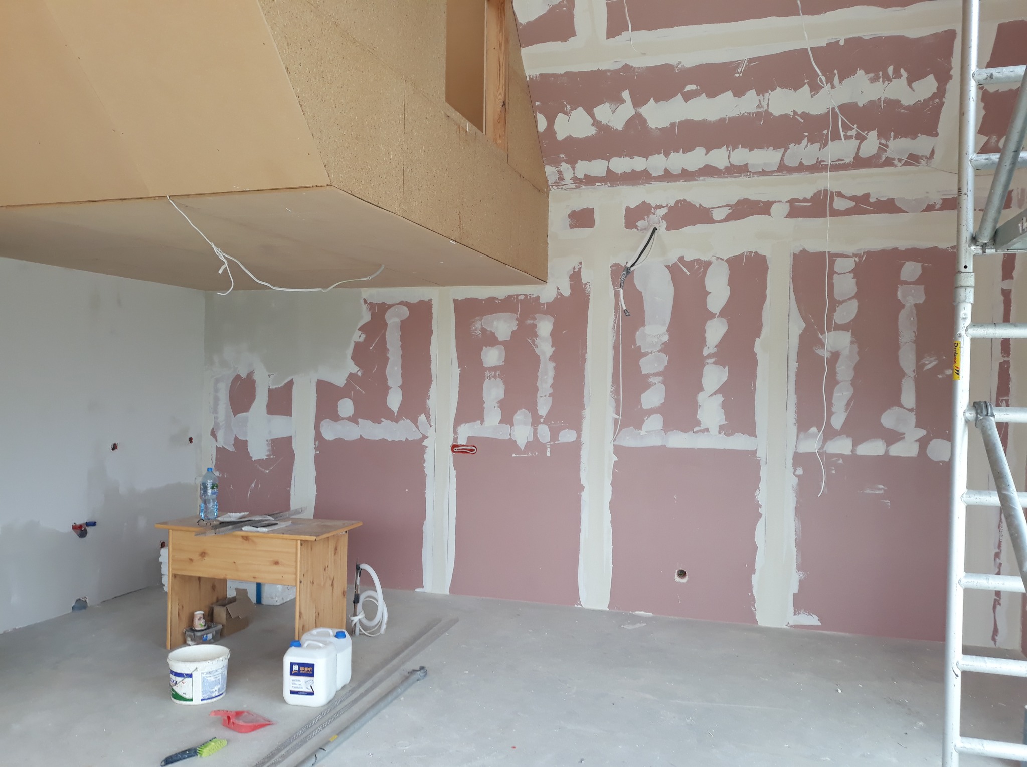 Wnętrze w trakcie remontu: ściany z płyt gipsowo-kartonowych, szpachlowane łączenia, drewniany stół roboczy, rusztowanie. Widoczne przewody elektryczne.