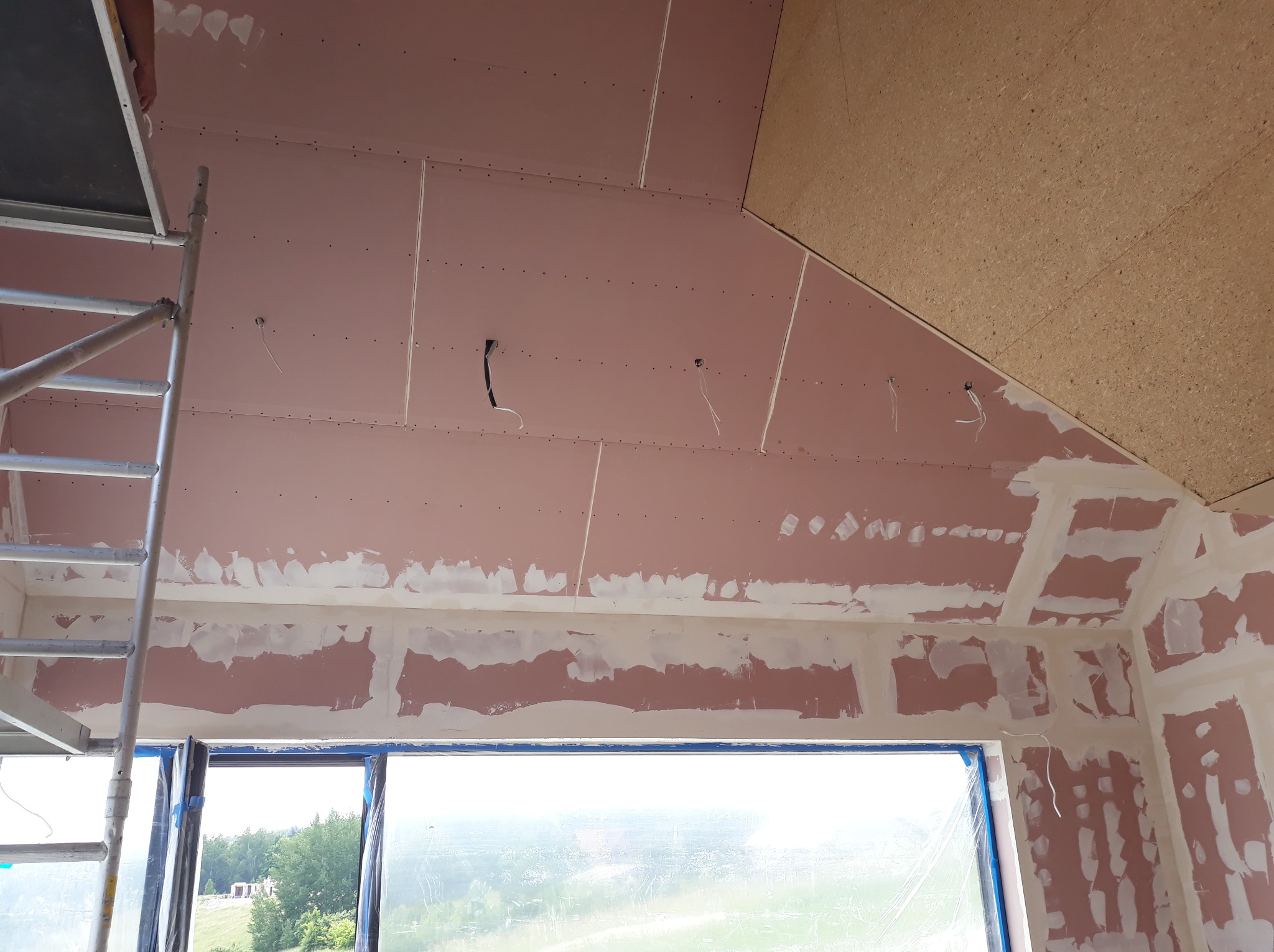 Poddasze w trakcie wykańczania: płyty gipsowe na skosach, częściowo zaszpachlowane łączenia, widoczne przewody elektryczne, rusztowanie i okno z widokiem na zieleń.