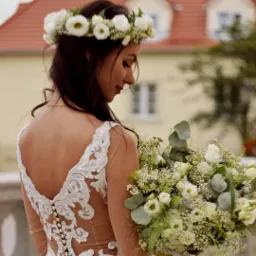 Panna młoda z ciemnymi włosami w koronie z białych kwiatów spogląda na bukiet ślubny z białych róż i zielonych dodatków na tle budynku z czerwoną dachówką.