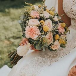 Pracownia Florystyczna Lawendowy kącik - Panna młoda trzyma bukiet ślubny z różowych i żółtych kwiatów, z łodygami owiniętymi kremową koronką, na tle sukni ślubnej z koronkowym wykończeniem.