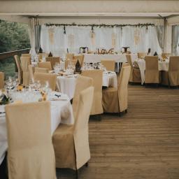 Pracownia Florystyczna Lawendowy kącik - Uroczysta dekoracja sali weselnej: okrągłe stoły z białymi obrusami, krzesła w beżowych pokrowcach, dekoracje ze łapaczami snów i napisem LOVE, drewniana podłoga.