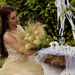 Pracownia Florystyczna Lawendowy kącik - Kobieta w złotej sukni trzyma bukiet z suszonych traw i kremowych róż, siedząc przy rustykalnym stole z napisem 'Dream' i kieliszkiem wody, w tle zielone krzewy i łapacz snów z białymi wstążkami.