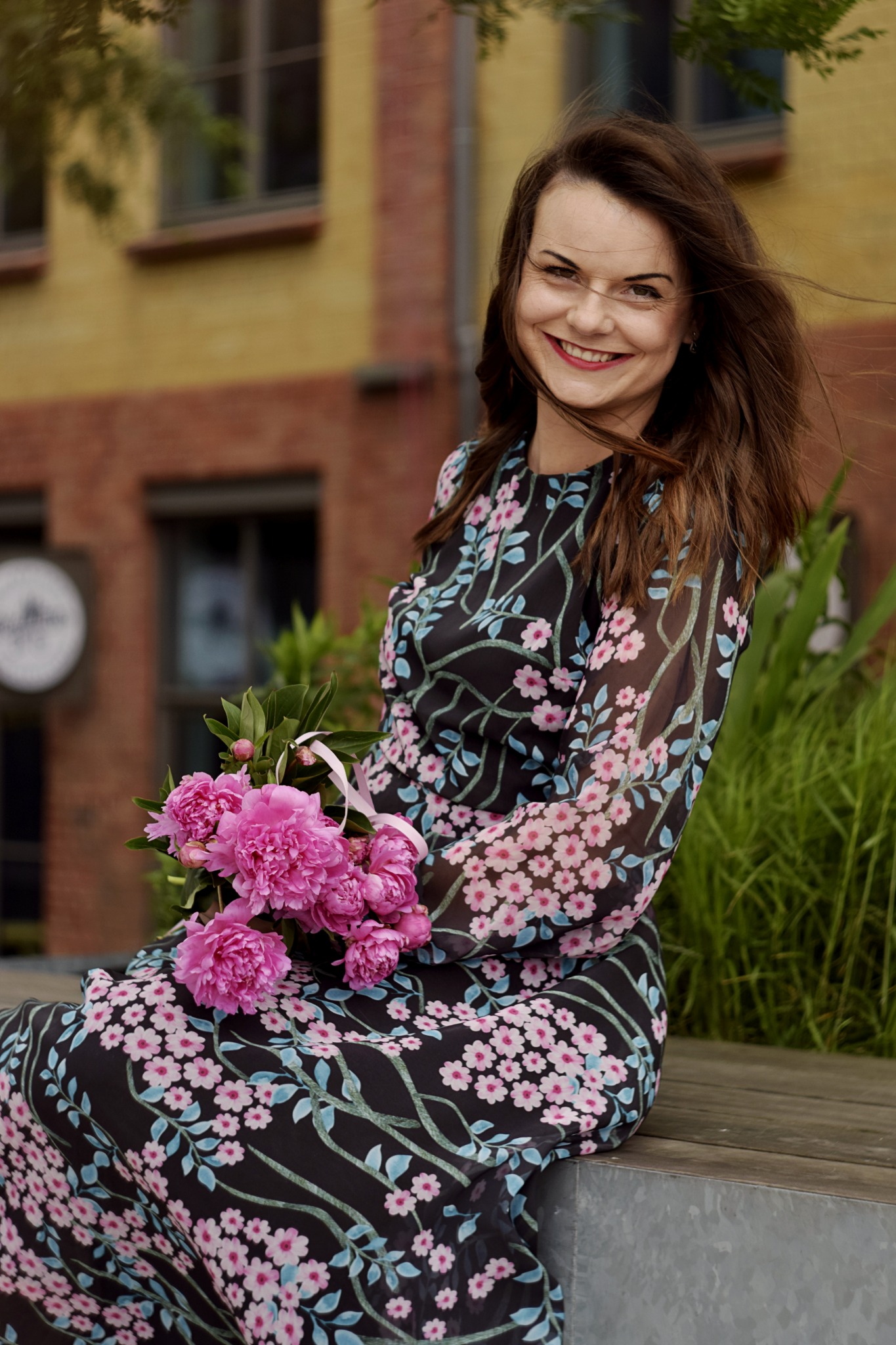 Uśmiechnięta kobieta w kwiecistej sukience siedzi na ławce, trzymając bukiet różowych piwonii z satynową wstążką, w tle ceglany budynek i zielona trawa.