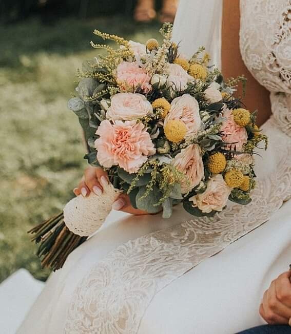 Panna młoda trzyma bukiet ślubny z różowych i żółtych kwiatów, z łodygami owiniętymi kremową koronką, na tle sukni ślubnej z koronkowym wykończeniem.