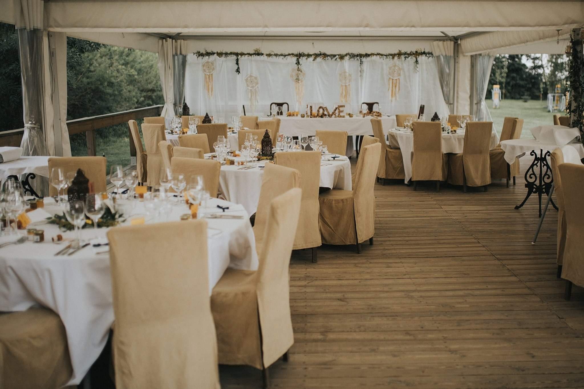 Uroczysta dekoracja sali weselnej: okrągłe stoły z białymi obrusami, krzesła w beżowych pokrowcach, dekoracje ze łapaczami snów i napisem LOVE, drewniana podłoga.