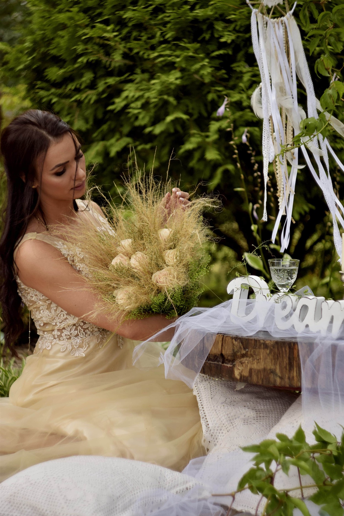 Kobieta w złotej sukni trzyma bukiet z suszonych traw i kremowych róż, siedząc przy rustykalnym stole z napisem 'Dream' i kieliszkiem wody, w tle zielone krzewy i łapacz snów z białymi wstążkami.