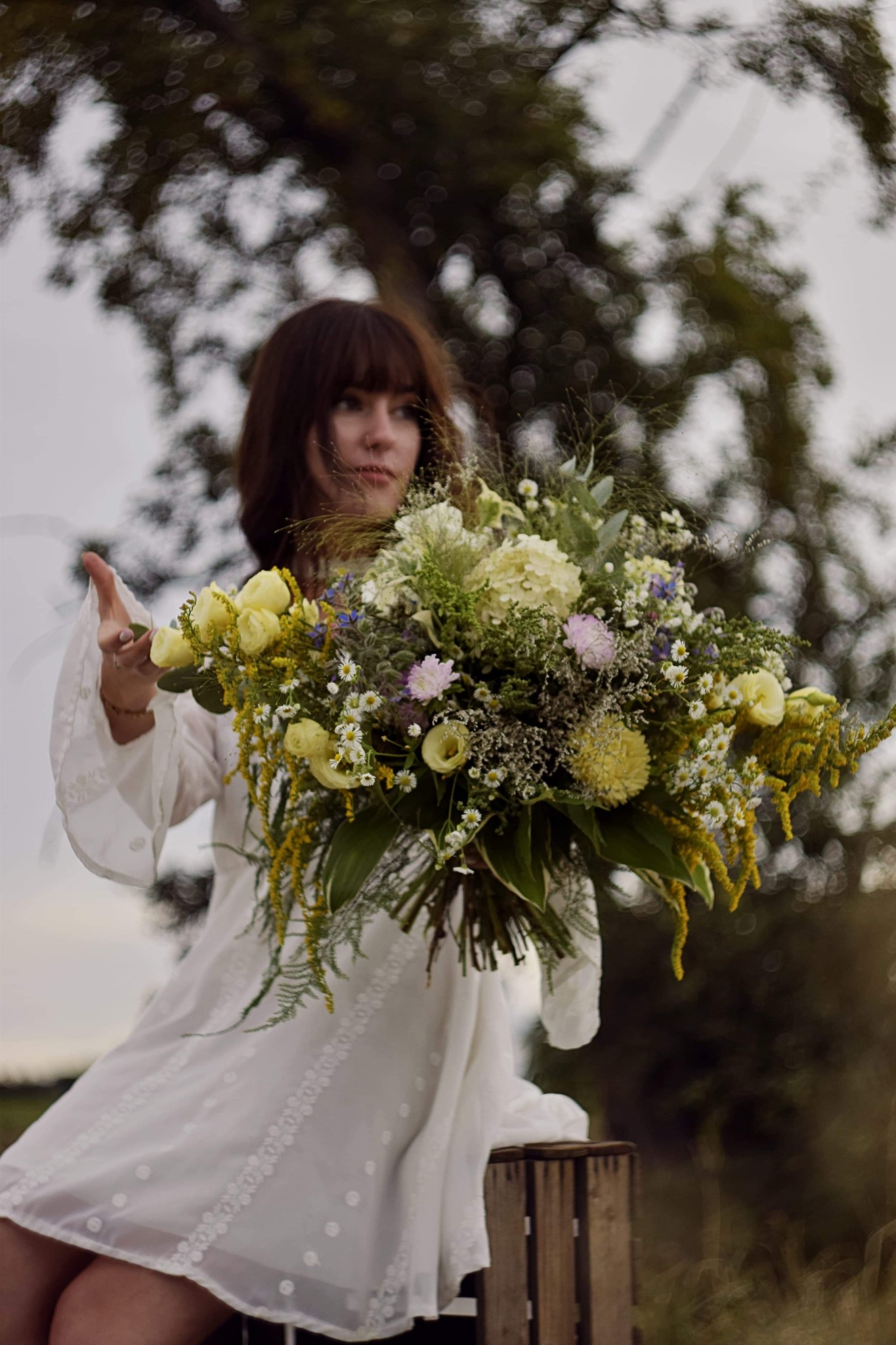 Kobieta w białej sukience trzyma duży bukiet mieszanych kwiatów w odcieniach żółci, bieli i fioletu na tle rozmytego drzewa.