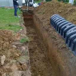 Wykopany wąski rów w ziemi z ułożonymi czarnymi elementami drenażowymi, operator mini koparki i mężczyzna z łopatą w tle, trawa i budynek w oddali.