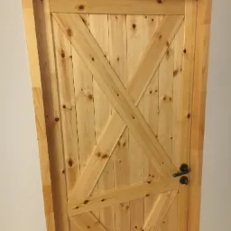 Drewniane drzwi wewnętrzne z surowego drewna sosnowego, zdobione krzyżującymi się listwami, z czarną klamką i zamkiem, zamontowane w ościeżnicy. Widoczna faktura drewna i sęki.