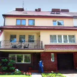 Dwupiętrowy dom po renowacji elewacji z balkonem i czerwonym dachem. Pracownik w niebieskim kombinezonie czyści elewację. Ogród z kwiatami i drabina obok garażu.