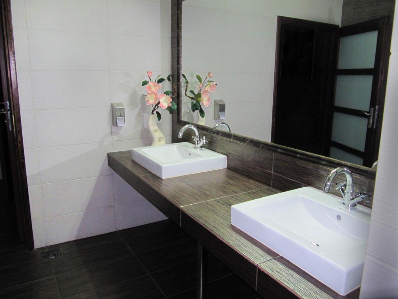 Nowoczesna łazienka z dwiema białymi umywalkami na blacie z płytek imitujących drewno, lustro z kwiatami i ciemne płytki podłogowe.