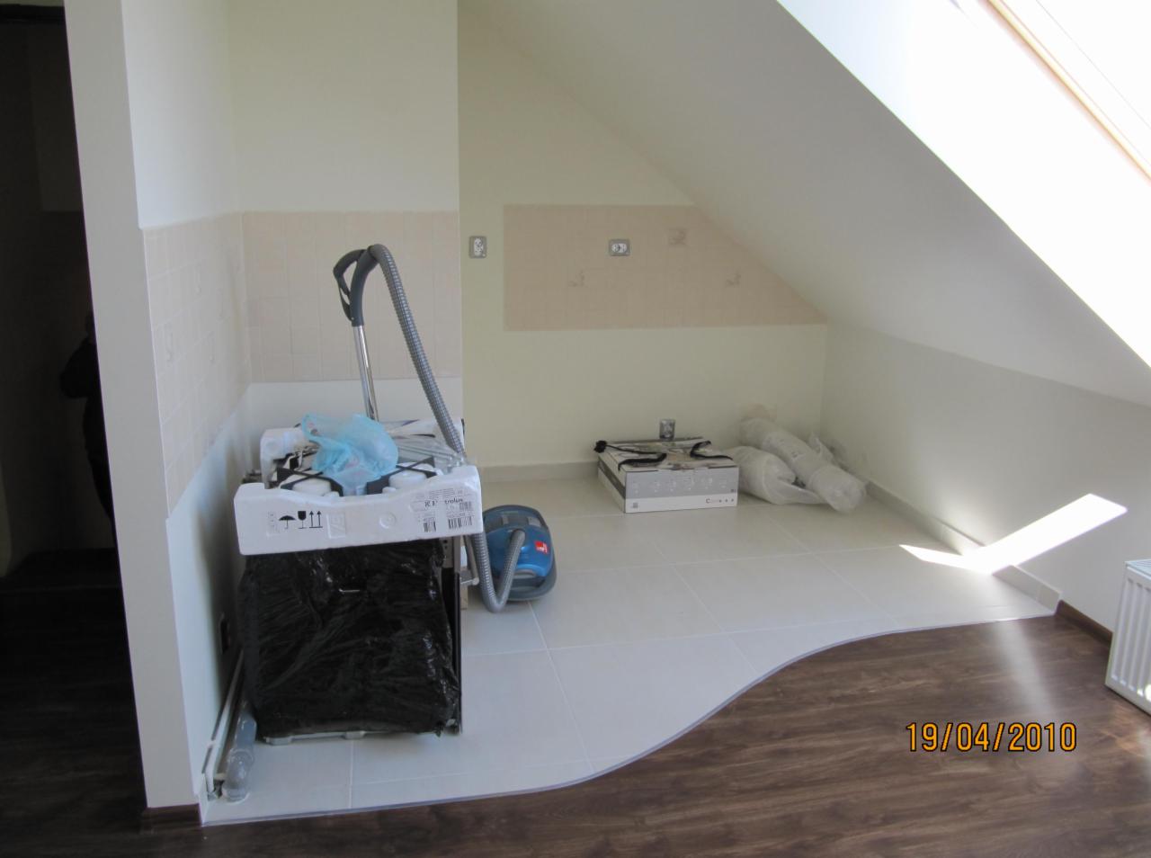 Poddasze po remoncie: białe płytki przechodzą w drewnopodobną podłogę. Nowa kuchenka i odkurzacz stoją na płytkach, ściany pomalowane na biało, okno dachowe.