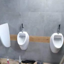 Trzy białe pisuary zamontowane na szarej ścianie z płytek, z widocznymi narzędziami i materiałami budowlanymi na podłodze.