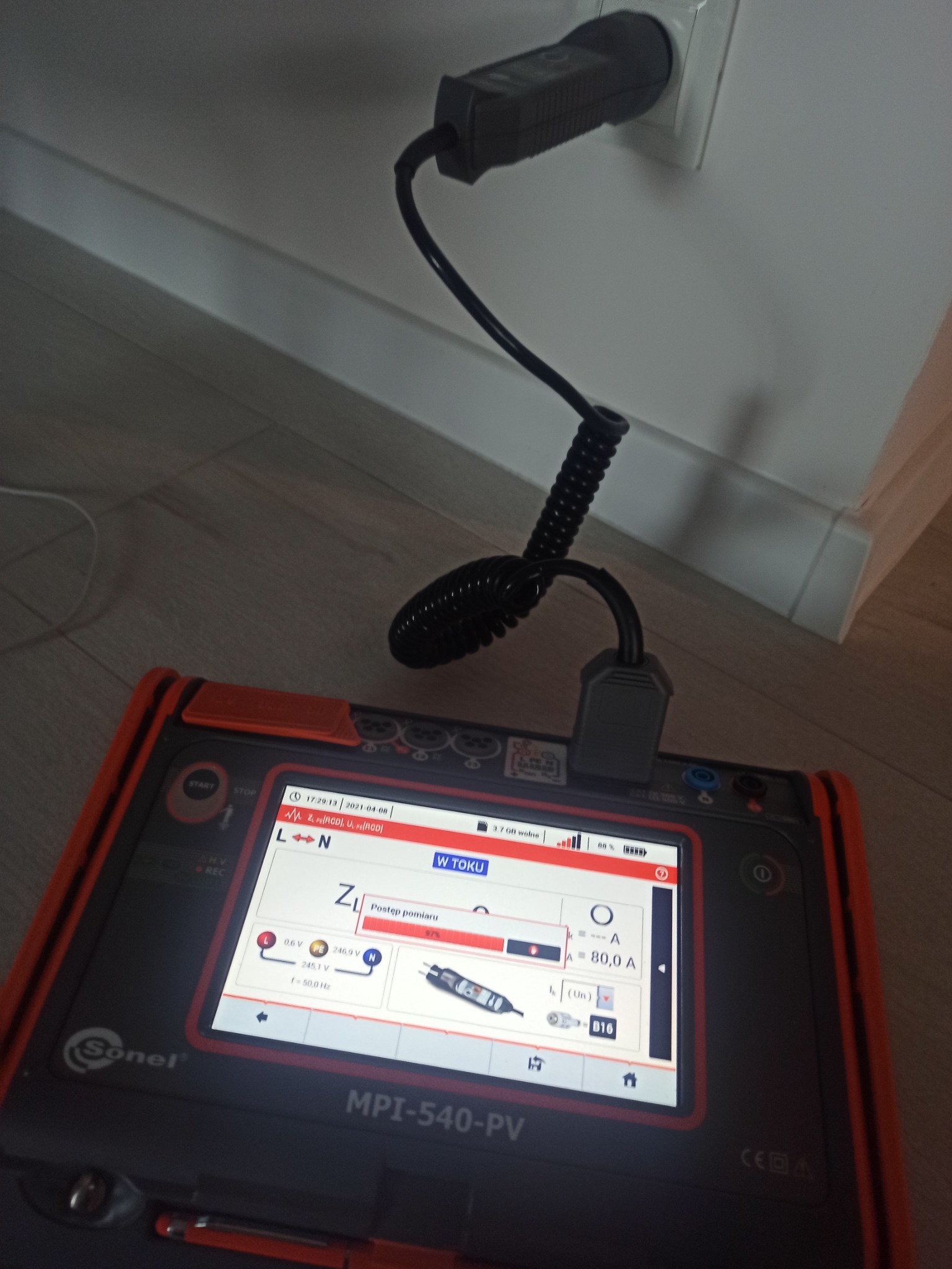 Miernik Sonel MPI-540-PV podłączony do gniazdka elektrycznego, wyświetlający postęp pomiaru impedancji pętli zwarcia na ekranie LCD.