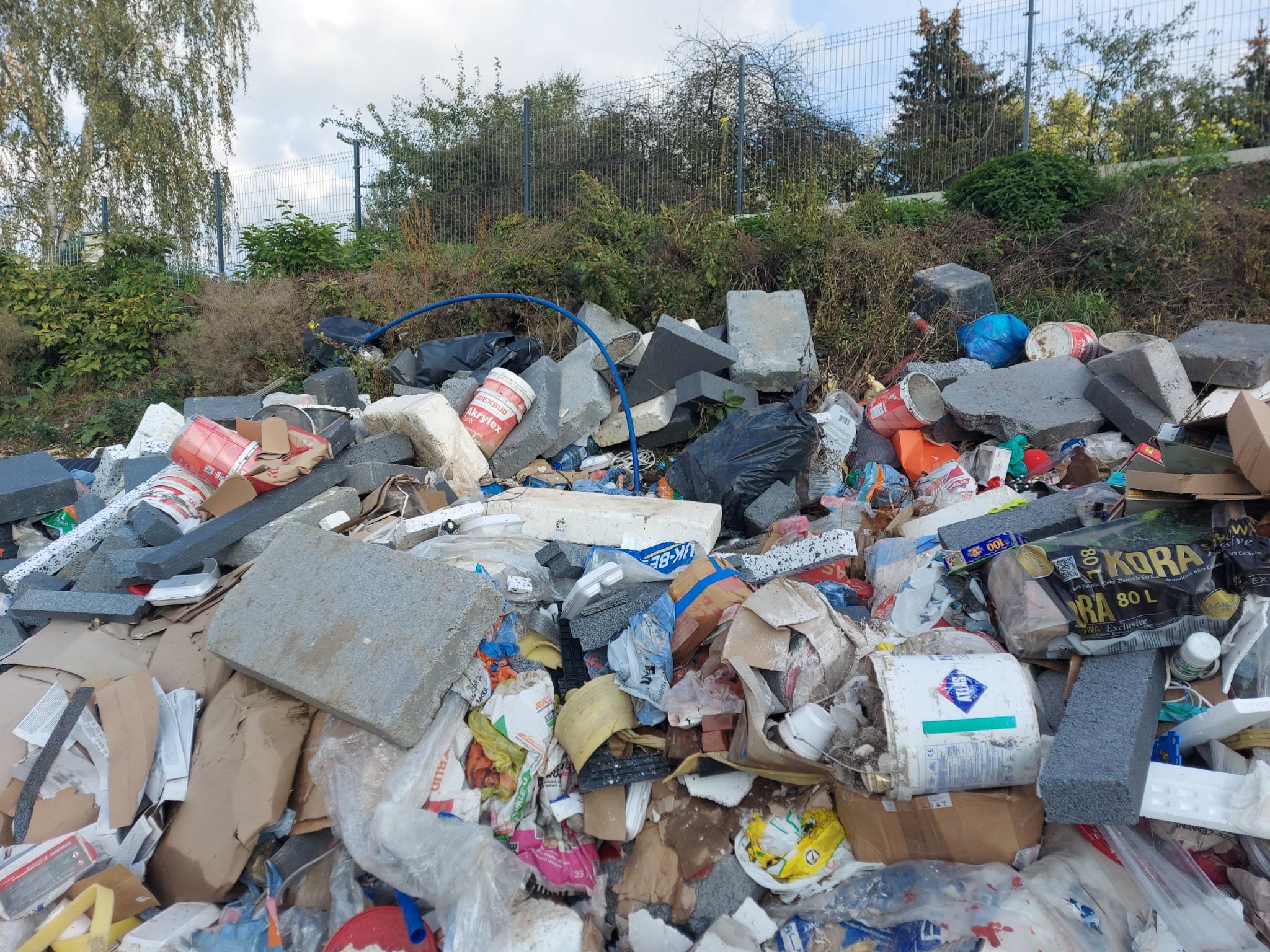 Stos gruzu, odpadów budowlanych i śmieci różnego rodzaju, w tym styropianu, kartonów i plastikowych opakowań, z widocznymi puszkami po farbach i wiaderkami, na tle zieleni i ogrodzenia.