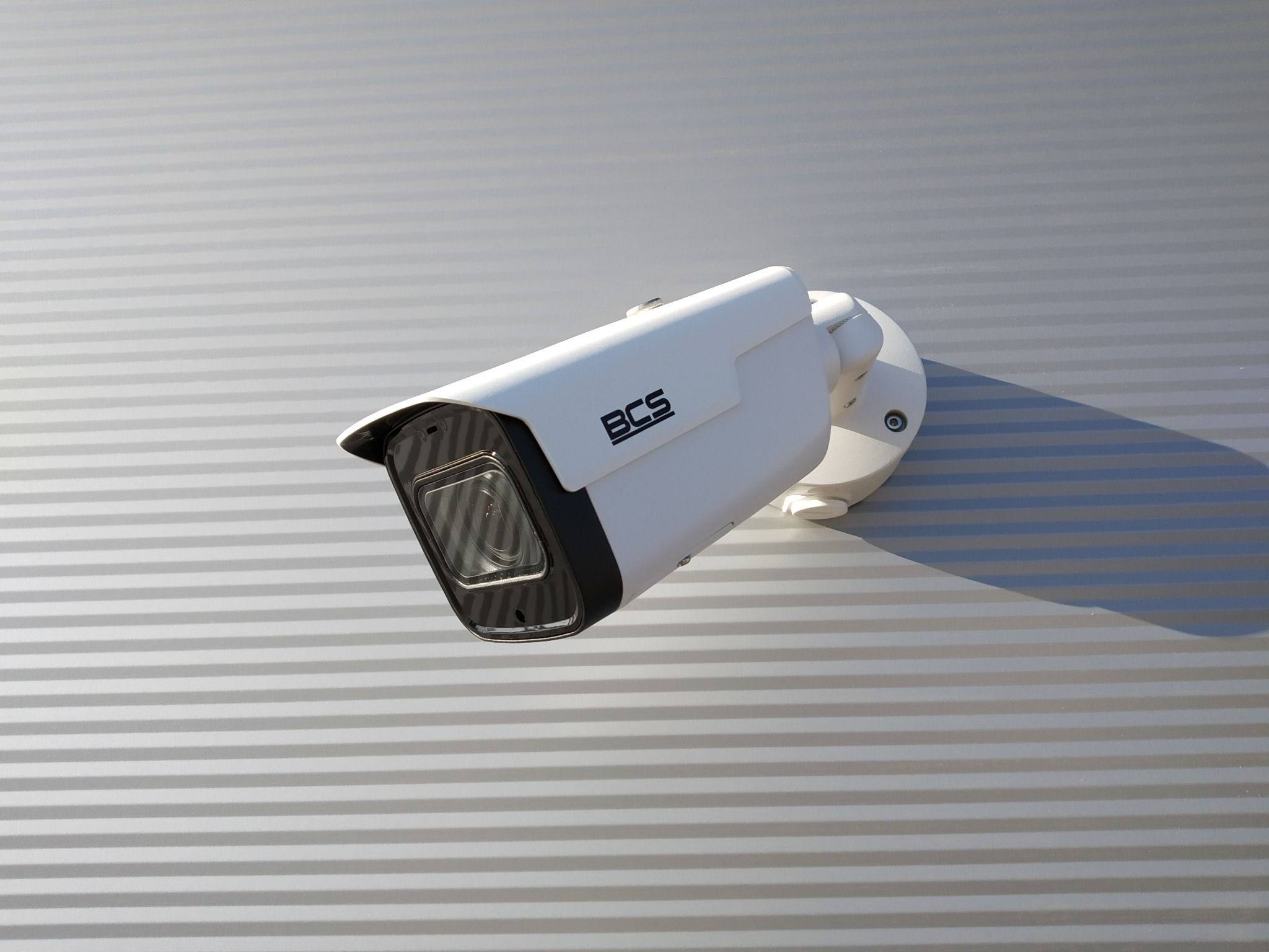 Biała kamera monitoringu BCS zamontowana na szarej, poziomej ścianie z widocznym cieniem od kamery.