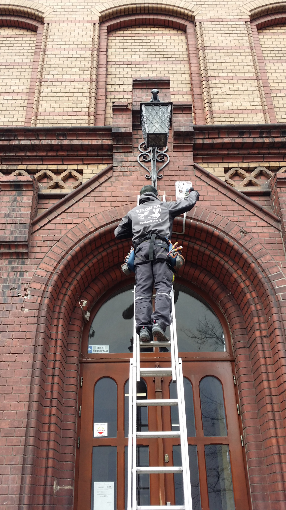 Pracownik na drabinie montuje urządzenie na elewacji budynku z czerwonej cegły, widoczny napis 'OBIEKT MONITOROWANY' na drzwiach.