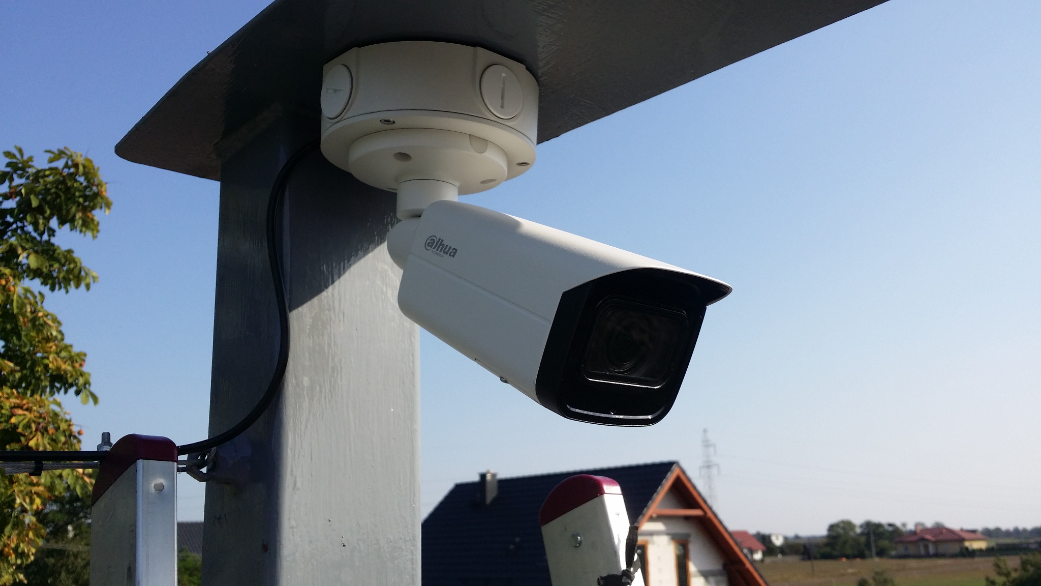 Kamera monitoringu Dahua zamontowana na szarej metalowej konstrukcji z widocznym kablem, w tle domy i niebieskie niebo.