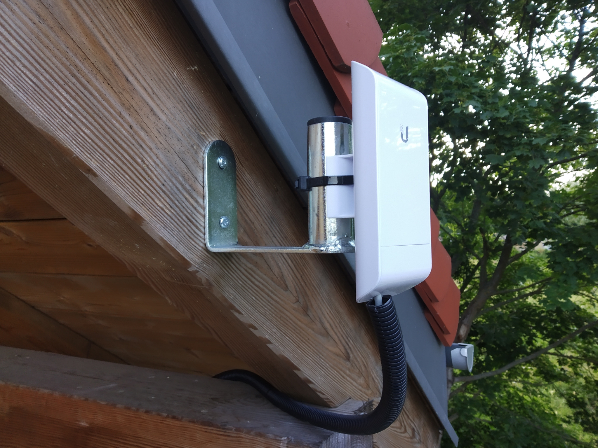 Zewnętrzny punkt dostępowy WiFi zamontowany na drewnianej elewacji domu, widoczny uchwyt montażowy i peszel z kablem, w tle drzewa.