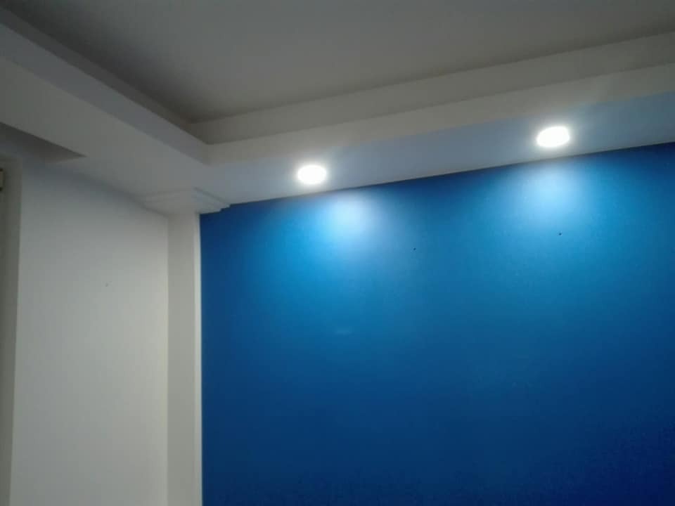 Pomieszczenie po odświeżeniu: jedna ściana w odcieniu głębokiego błękitu, oświetlona dwoma punktowymi lampami sufitowymi, pozostałe ściany białe, widoczny fragment listwy przysufitowej.