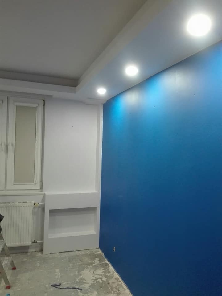 Świeżo pomalowana na niebiesko ściana w pokoju z białymi elementami wykończeniowymi i zamontowanym oświetleniem sufitowym, widoczny fragment drabiny malarskiej.