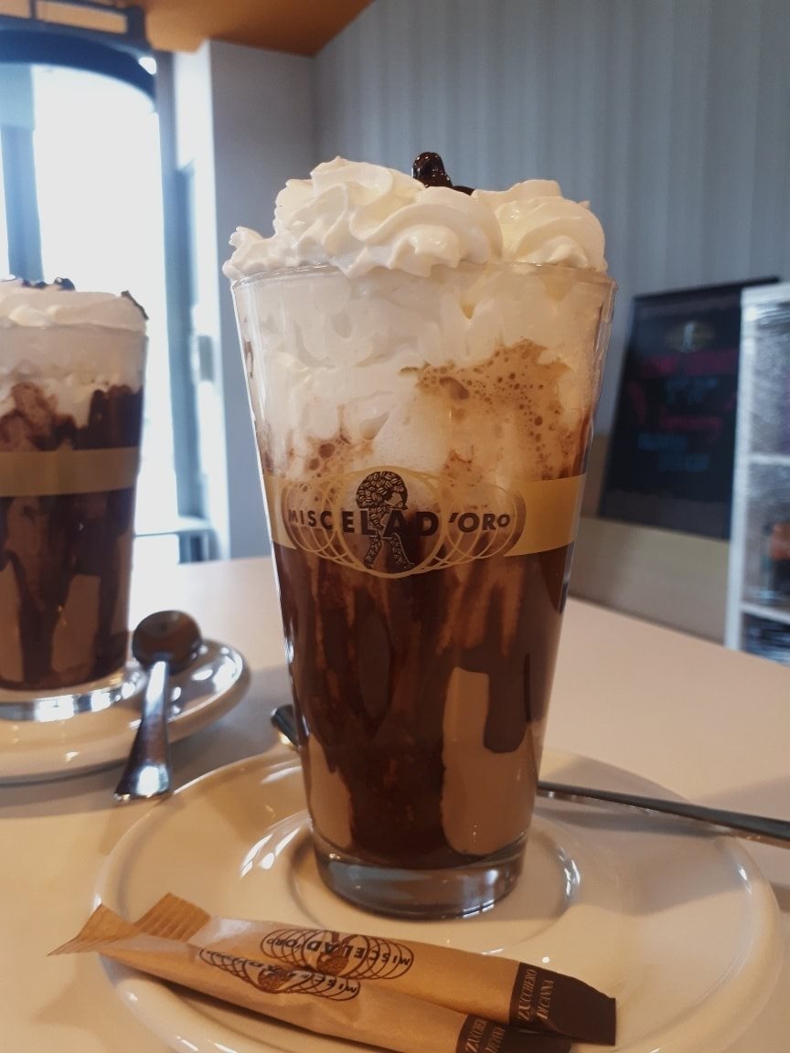 Szklanka kawy mrożonej z bitą śmietaną i polewą czekoladową na białym talerzyku, obok dwa opakowania cukru z logo marki kawy.