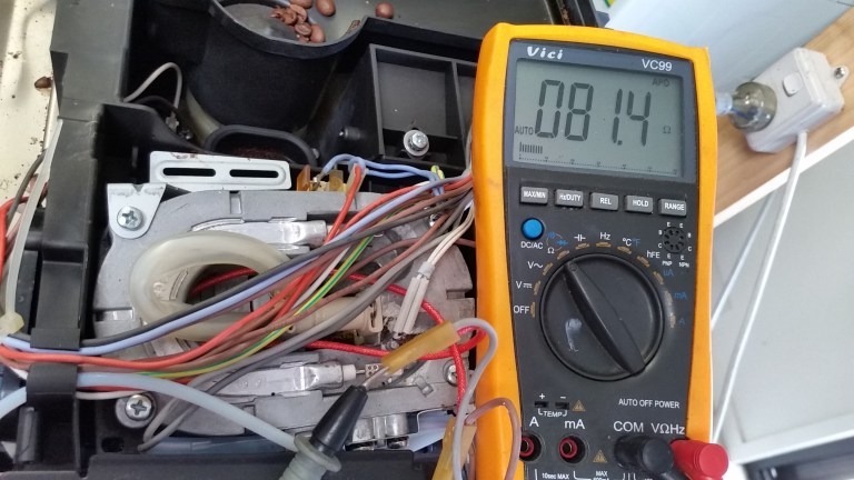 Wnętrze otwartego ekspresu do kawy z widocznymi przewodami i elementami elektronicznymi, obok żółty multimetr wskazujący 81.4 omów, prawdopodobnie podczas pomiaru rezystancji grzałki.