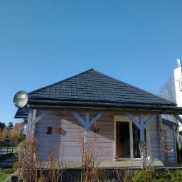 Niski, parterowy budynek z szarą elewacją, ciemnym dachem z dachówki i biało malowanymi drewnianymi elementami konstrukcyjnymi, widoczna antena satelitarna na dachu i dekoracje na ścianie, otoczony...