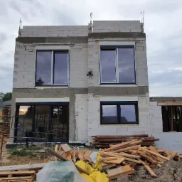 Dwukondygnacyjny budynek w stanie surowym otwartym z zamontowanymi oknami, widoczne materiały budowlane Ytong i sterta drewna na placu budowy.