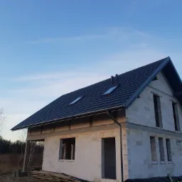 Surowy stan domu jednorodzinnego w trakcie budowy, z widocznym ciemnym dachem z dwoma oknami dachowymi i zamontowanymi rynnami, ściany z białych bloczków, niezadaszony ganek, w tle drzewa...