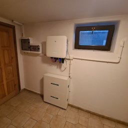 freeDOM fotowoltaika Marcin Szałacki - Kompletna instalacja fotowoltaiczna w piwnicy: inwerter GoodWe, dwuczęściowy magazyn energii, rozdzielnia elektryczna i okno piwniczne w tle.