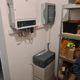 freeDOM fotowoltaika Marcin Szałacki - Instalacja domowa: biały inwerter, szara skrzynka bezpieczników i dwuczęściowy magazyn energii Pylontech na tle ściany i metalowego regału.