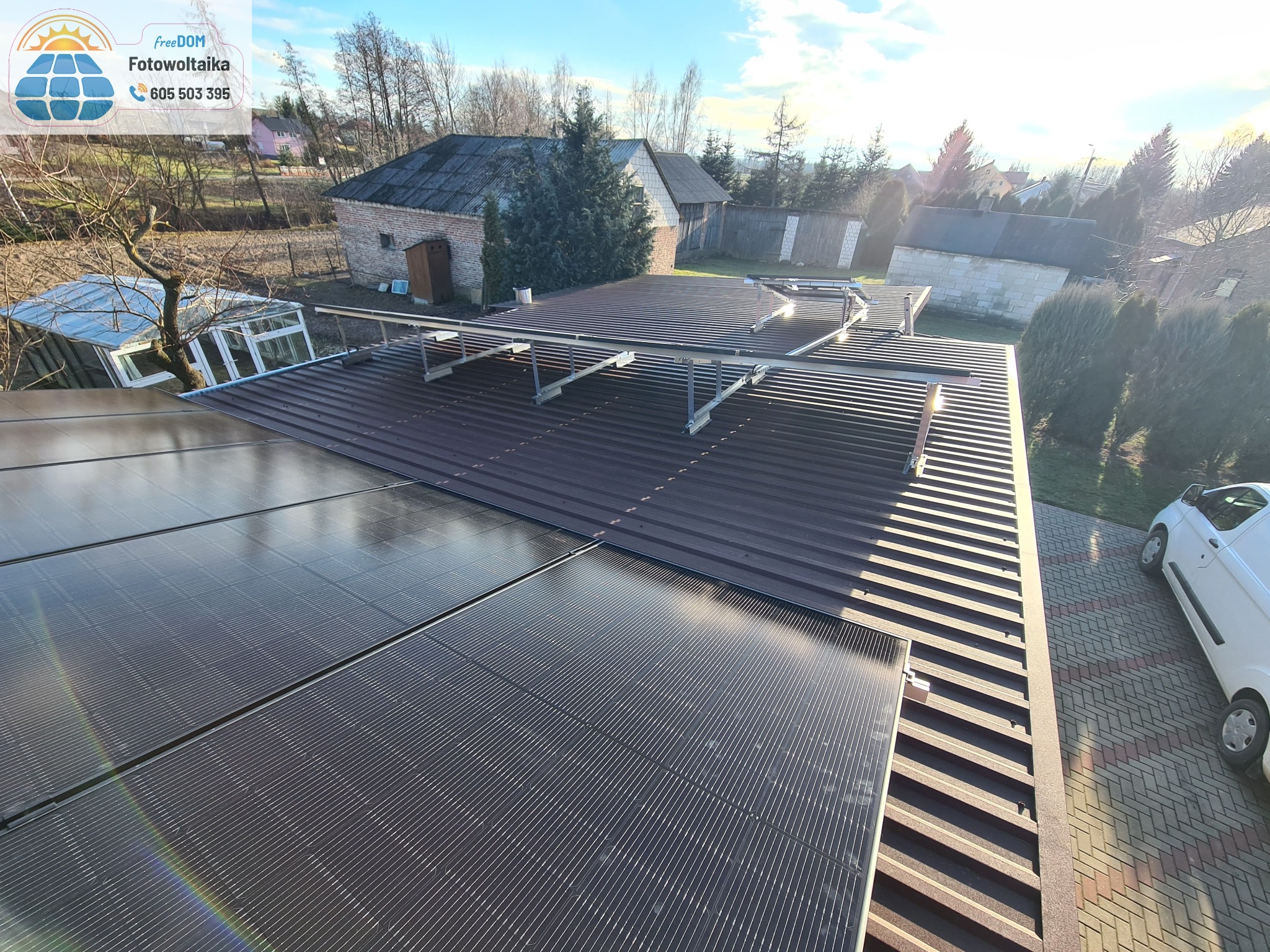 Montaż paneli słonecznych na brązowym dachu blaszanej konstrukcji z widocznymi elementami mocującymi, w tle zabudowa mieszkalna i błękitne niebo.