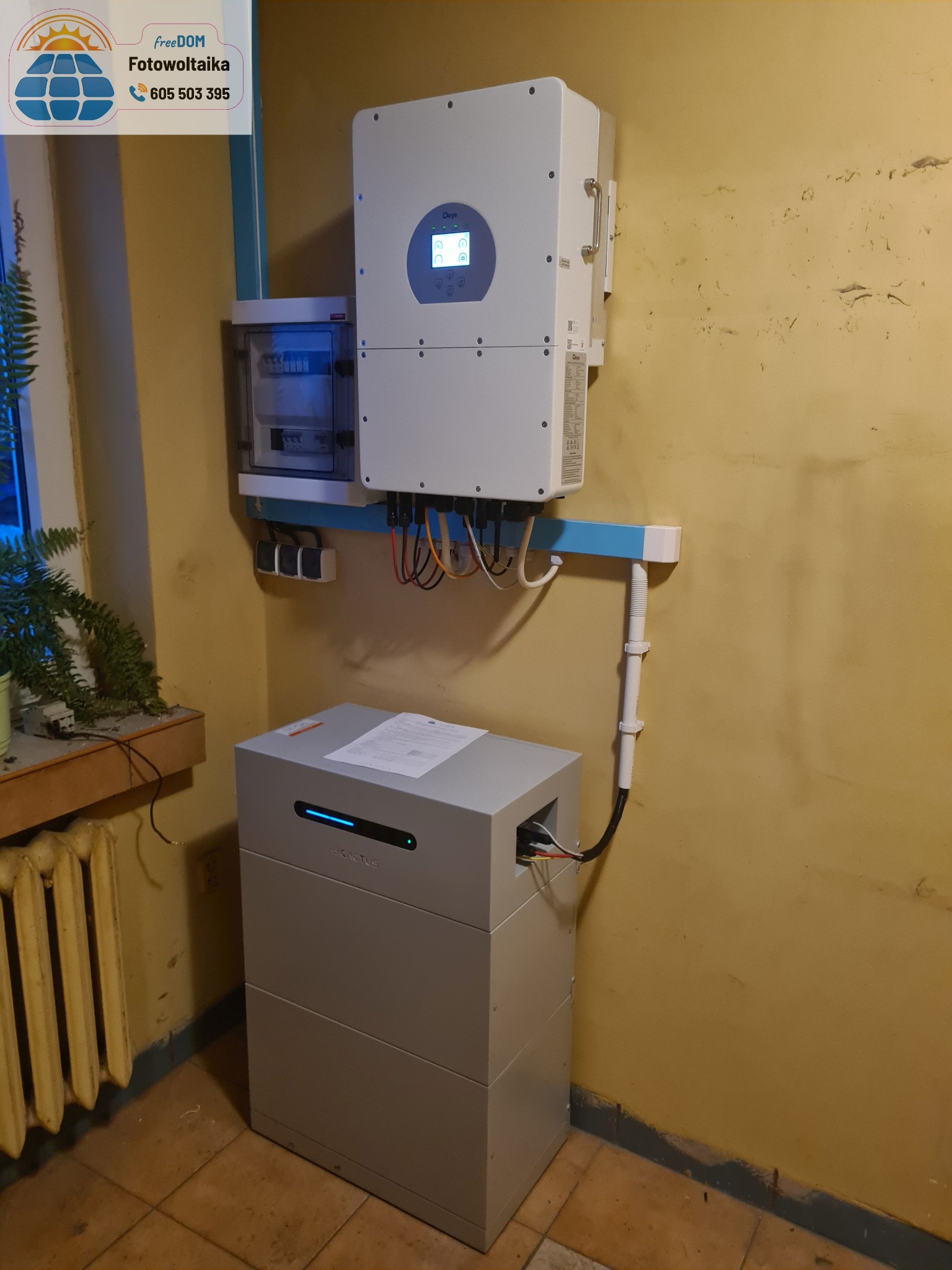 Szara bateria i biały inwerter zamontowane na żółtej ścianie wewnątrz budynku. Widoczne przewody i instalacja elektryczna. Obok okno z rośliną.