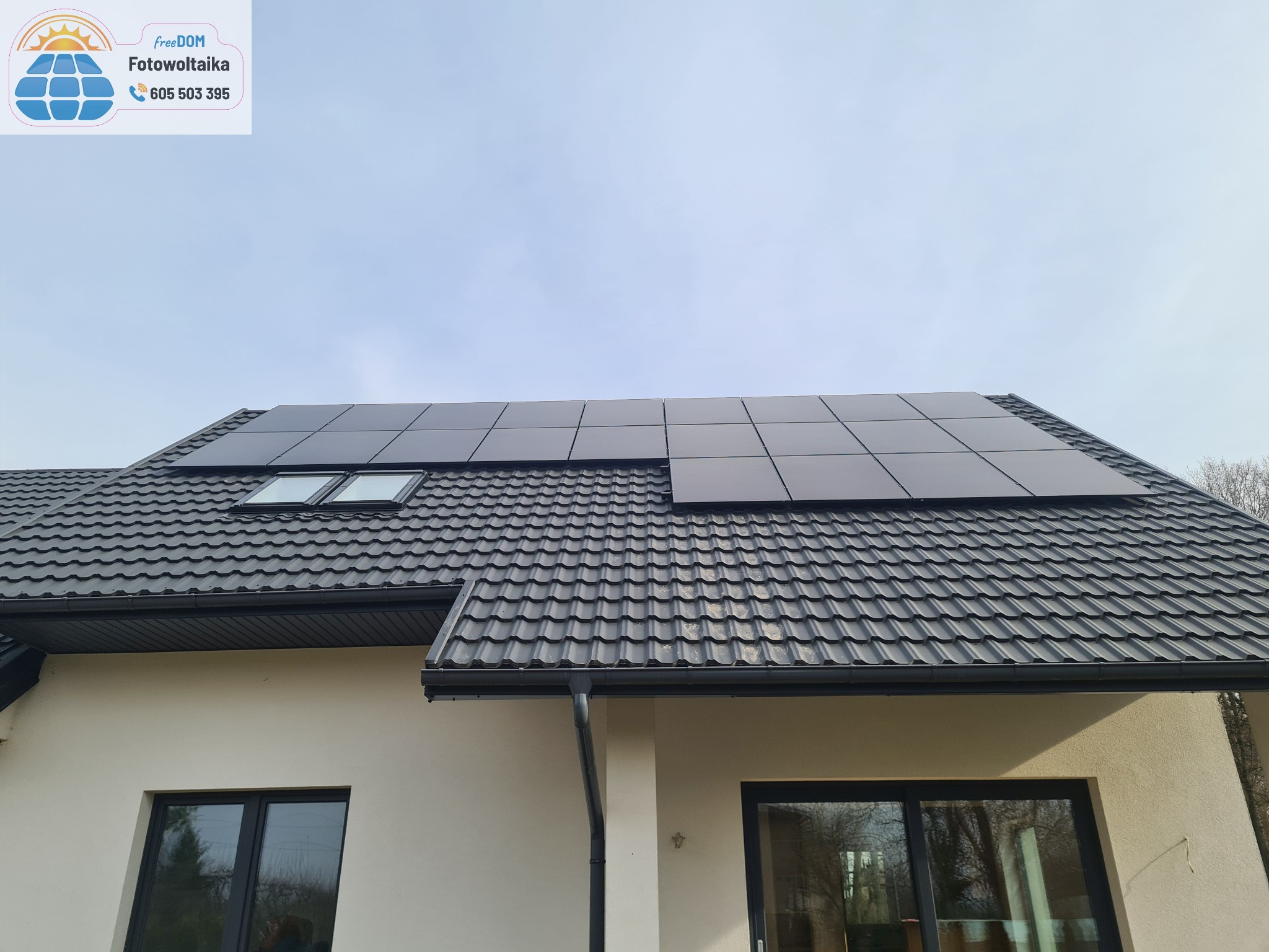 Panele słoneczne na dachu domu z dachówką w kolorze antracytu i oknami dachowymi. Widok od dołu, błękitne niebo w tle.