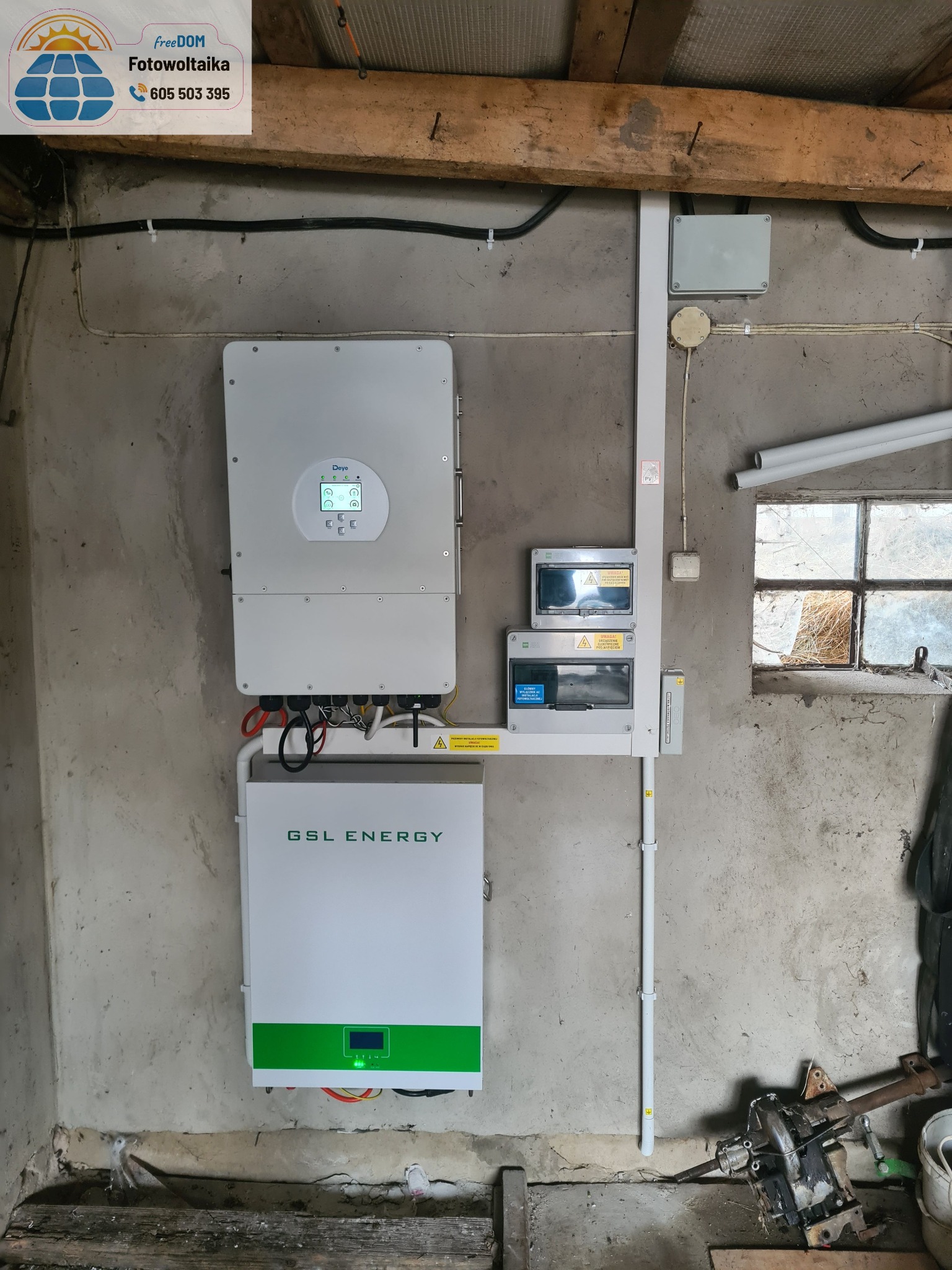 Zamontowany inwerter i magazyn energii GSL w szarej kotłowni z oknem. Instalacja w rurkach, tablica rozdzielcza. Widoczne przewody i elementy montażowe.