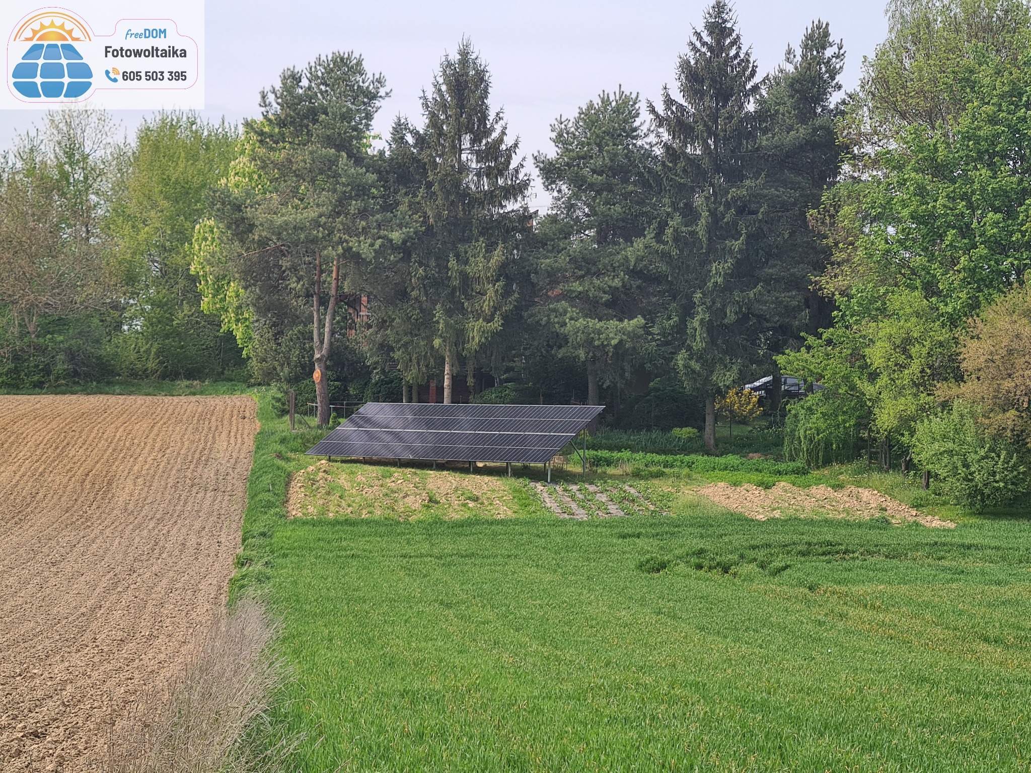 Panel fotowoltaiczny na skraju pola uprawnego, otoczony zielenią drzew. Widoczne plony rolne i logo firmy w górnym rogu.