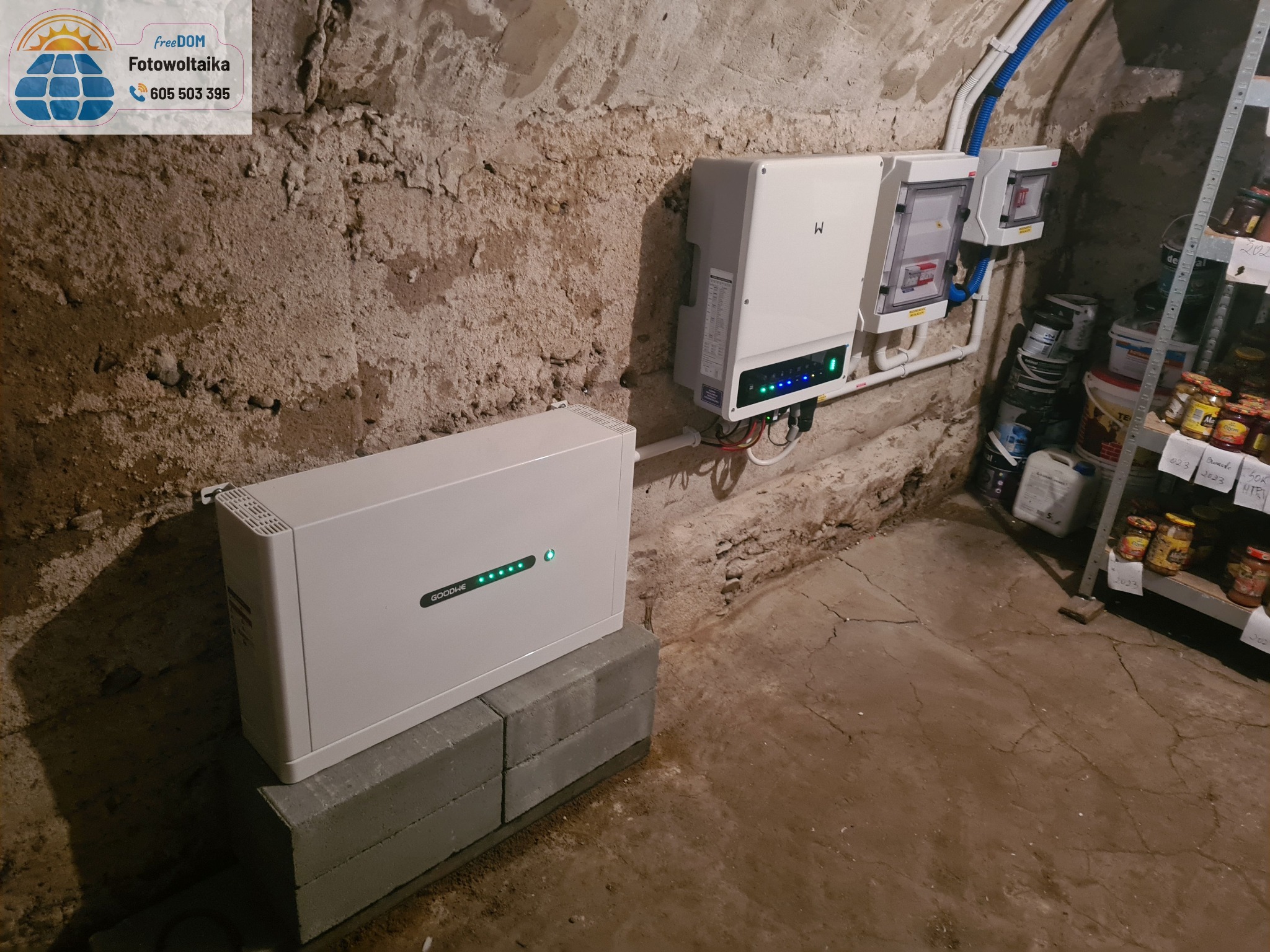 Instalacja magazynu energii GoodWe w piwnicy o surowych ścianach. Widoczny inwerter, zabezpieczenia i półka z przetworami. Realizacja w trudnych warunkach.