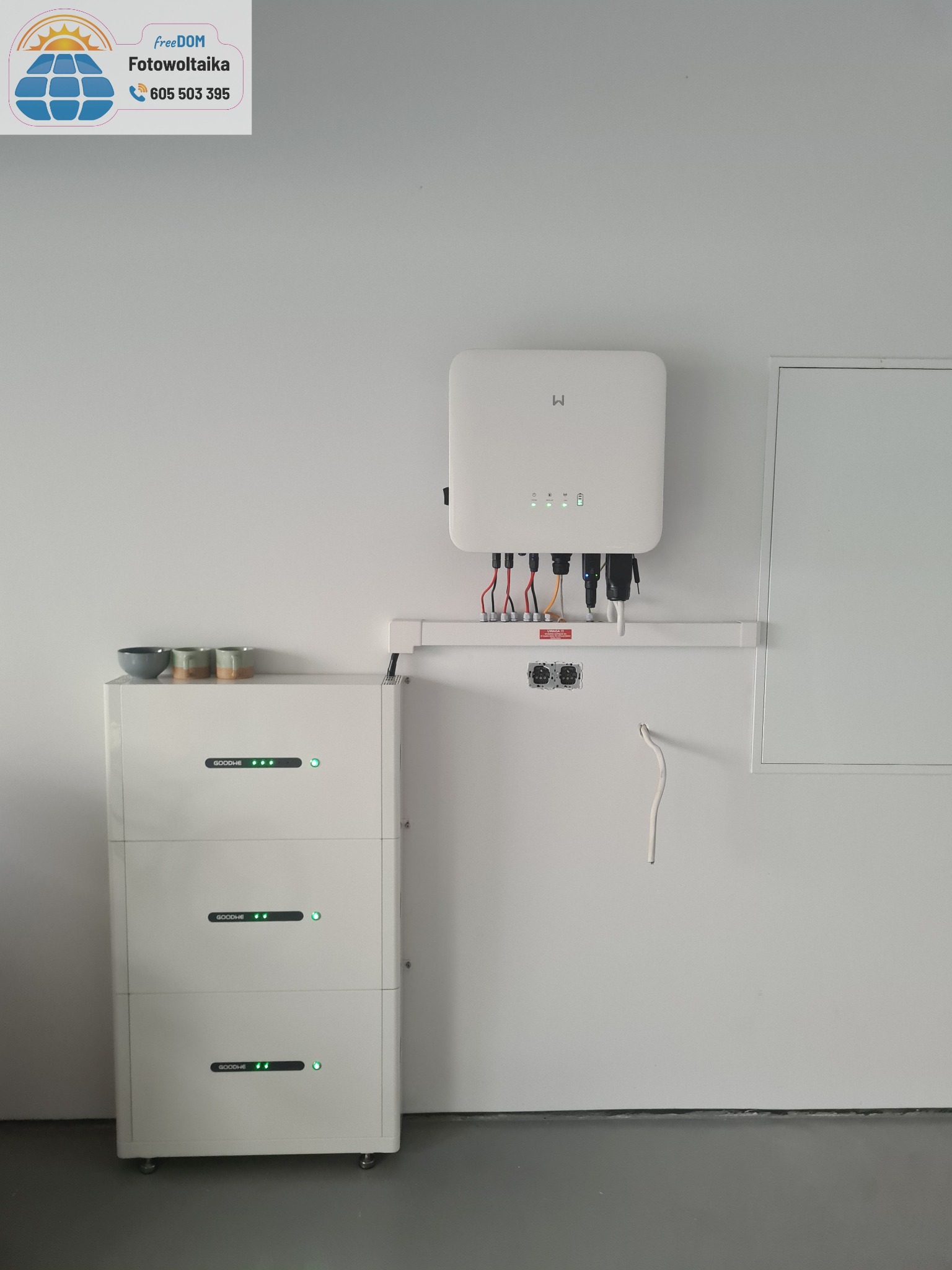 Biały magazyn energii GoodWe z trzema modułami i falownik na ścianie, kable i rury. Minimalistyczne wnętrze. Na górze magazynu stoją trzy małe miseczki.