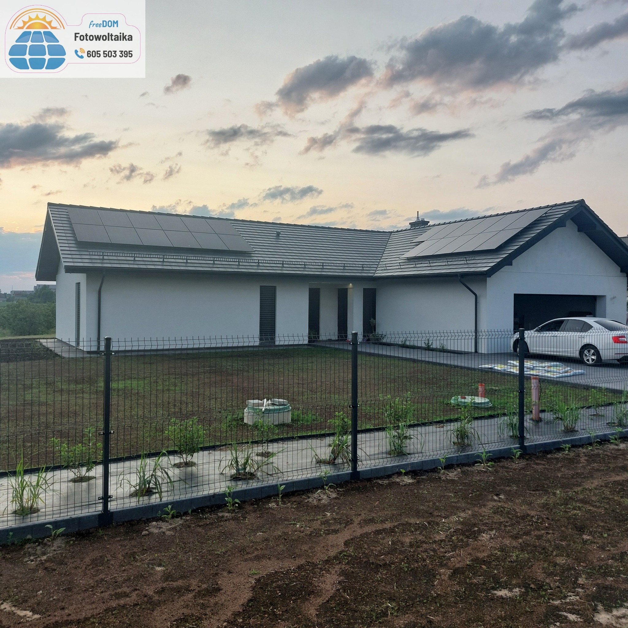 Nowoczesny dom jednorodzinny z panelami słonecznymi na dachu, widoczny zza ogrodzenia. W tle pochmurne niebo.
