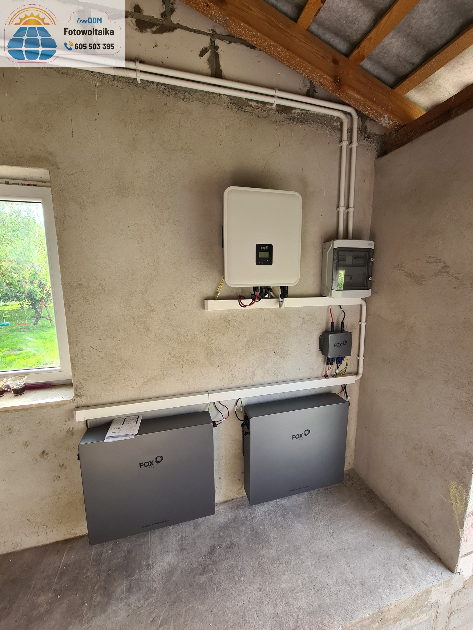 Instalacja magazynu energii FOX-ESS z inwerterem w surowym wnętrzu. Widoczne kable i rurki instalacyjne. Na podłodze leżą instrukcje. Widok z okna na zieleń.