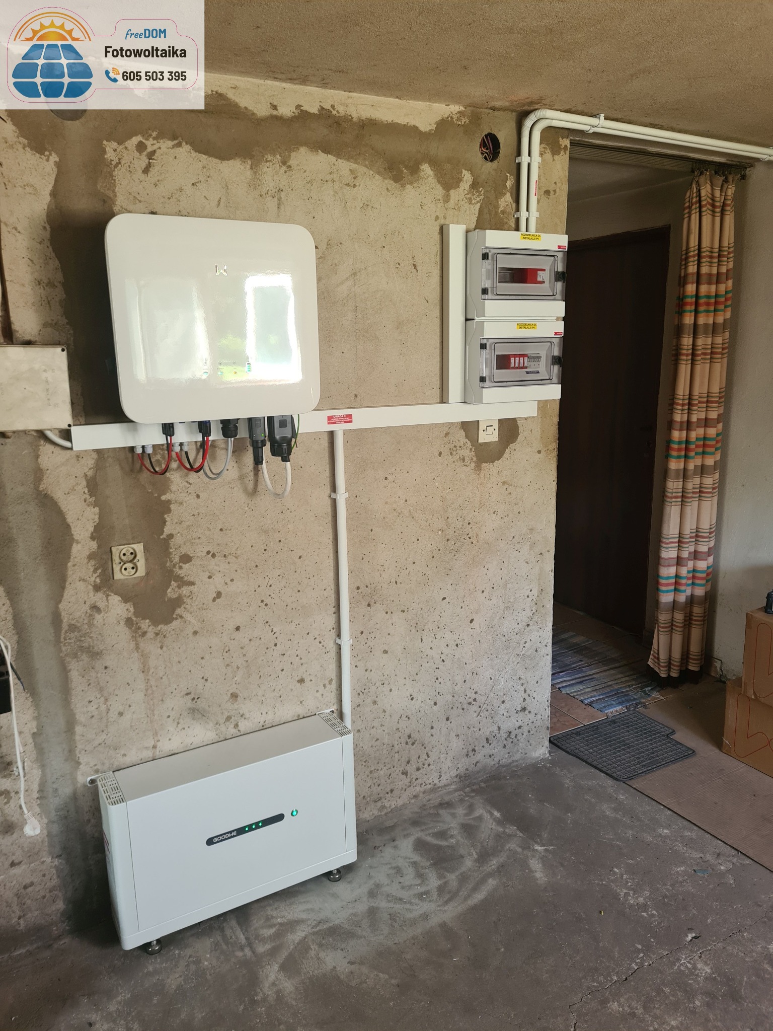 Zainstalowany magazyn energii Goodwe z inwerterem na ścianie o surowym wykończeniu, obok rozdzielnica elektryczna. Widoczne przewody i rury instalacyjne.