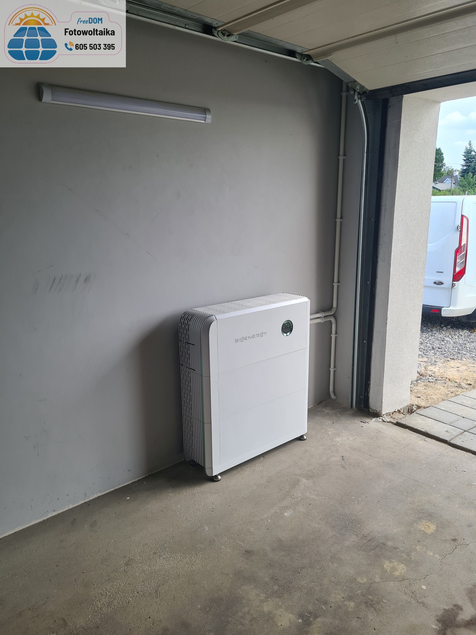 Biały magazyn energii Eigenergy w garażu, widoczny fragment białego busa na zewnątrz. Szara ściana, betonowa podłoga, oświetlenie LED.