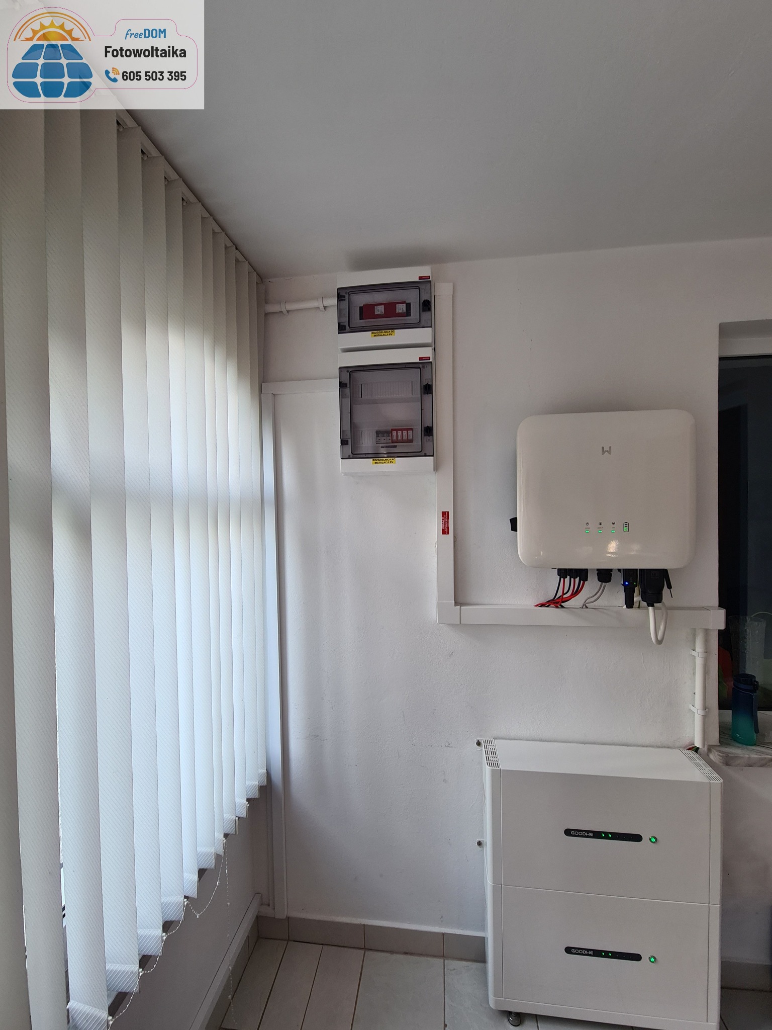 Wnętrze z zamontowanym magazynem energii GoodWe, inwerterem i rozdzielnicą. Widoczne białe żaluzje pionowe i szara płytka podłogowa. Całość w jasnej tonacji.