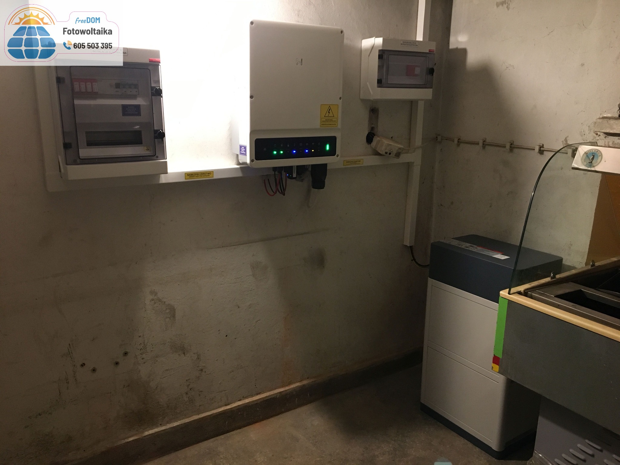 Instalacja elektryczna: rozdzielnice, falownik z sygnalizacją LED i akumulator w pomieszczeniu o surowych ścianach. Elementy instalacji fotowoltaicznej.