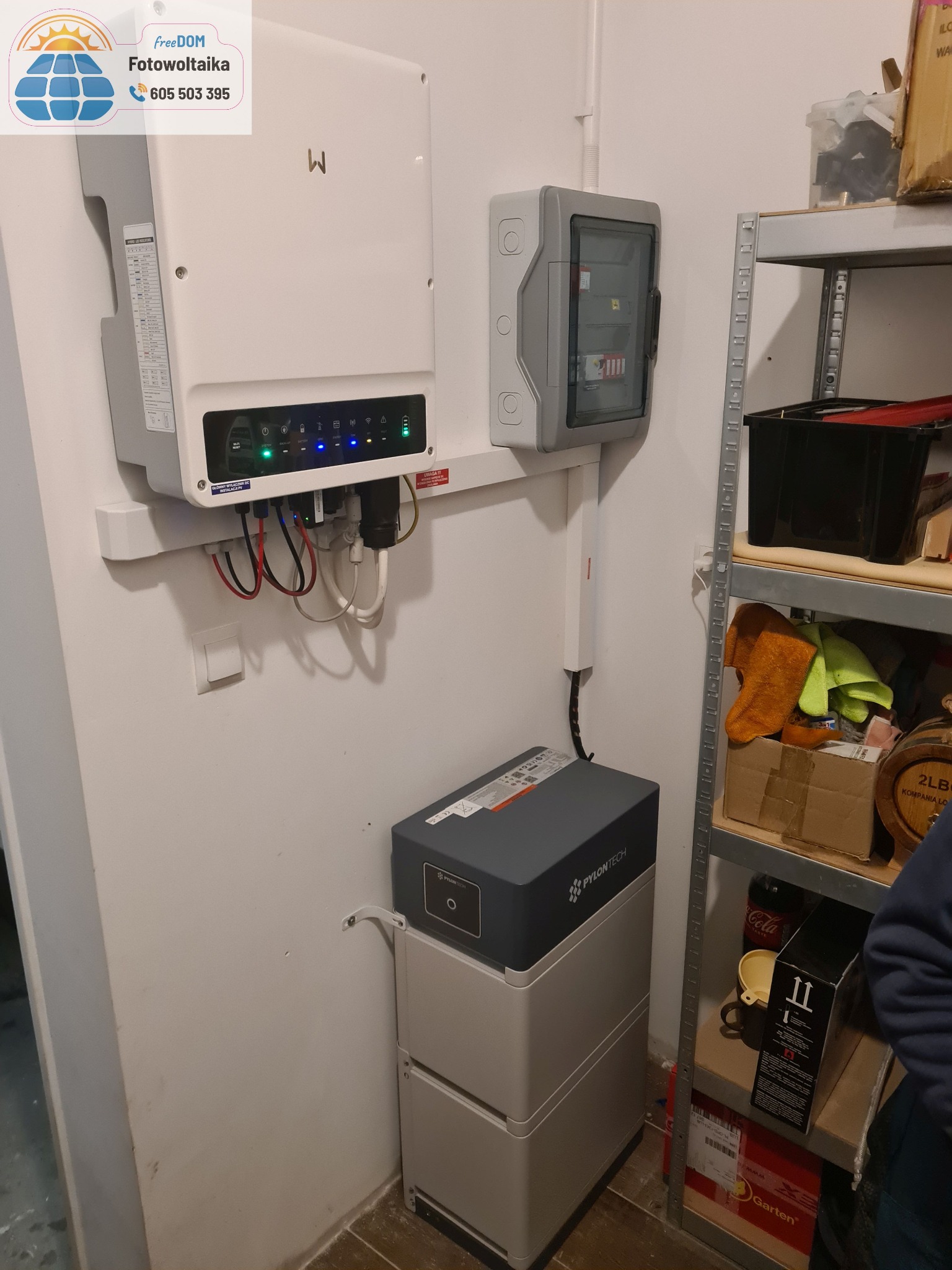 Instalacja domowa: biały inwerter, szara skrzynka bezpieczników i dwuczęściowy magazyn energii Pylontech na tle ściany i metalowego regału.