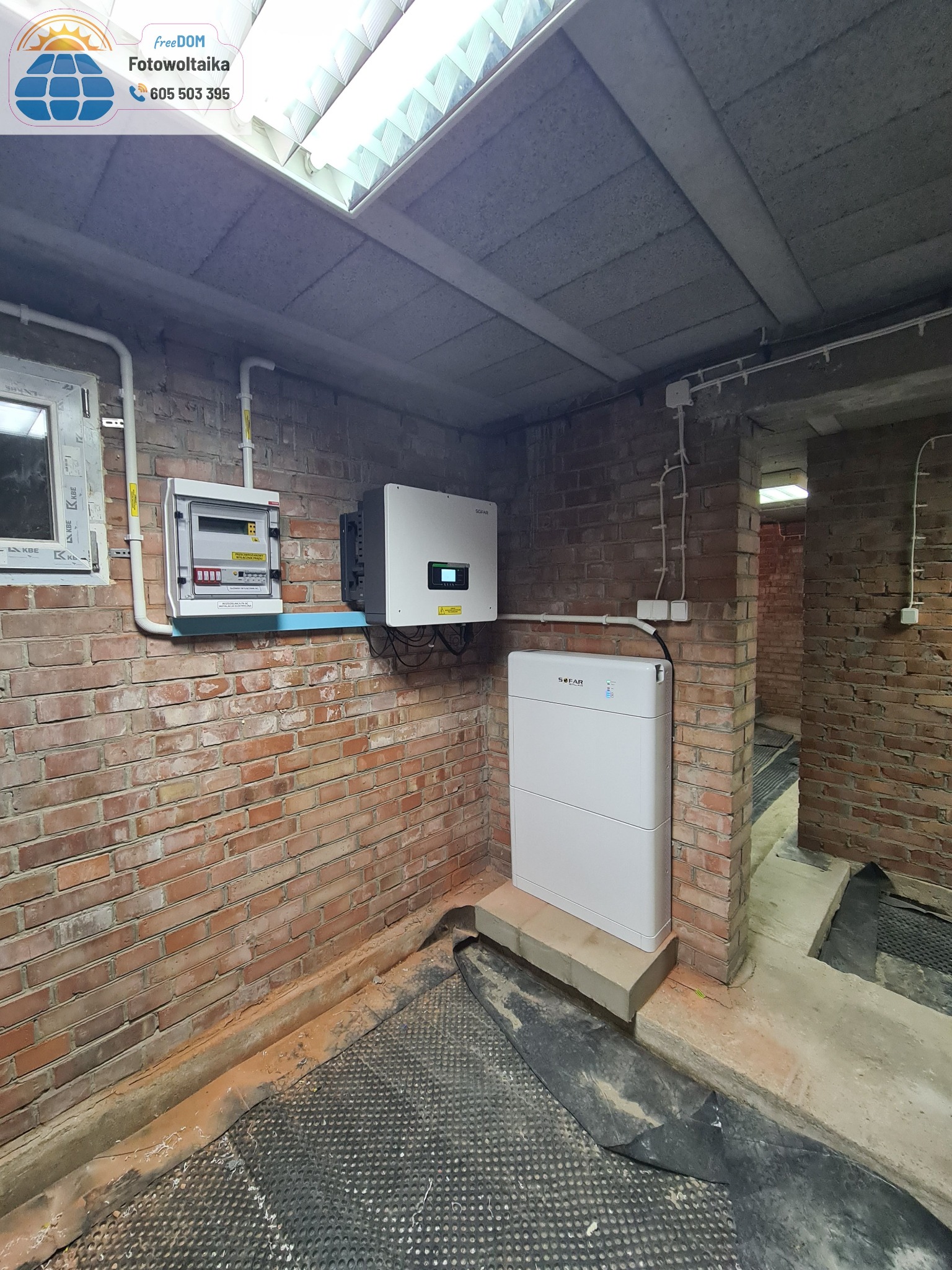 Instalacja magazynu energii SOFAR z rozdzielnicą i inwerterem na ceglanej ścianie w piwnicy. Widoczne kable i rury instalacyjne.